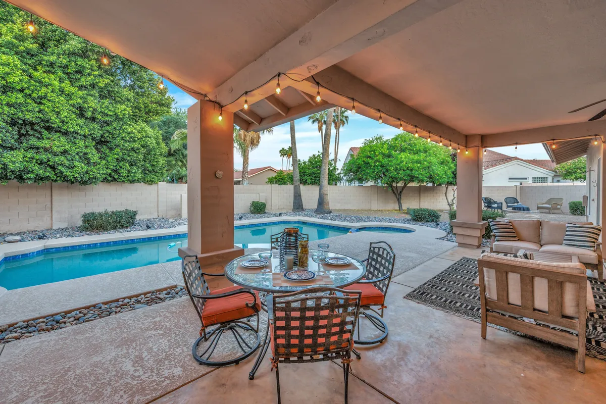 This image features a beautifully landscaped backyard with a serene swimming pool, a spacious dining table, and comfortable seating area. String lights overhead add ambiance, while lush greenery and open skies emphasize the inviting outdoor lifestyle. The scene captures sunset views and tranquil moments by the pool, suggesting an ideal spot for relaxation or winding down after work.