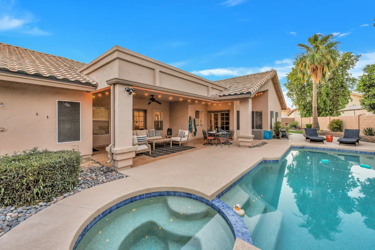 A beautiful backyard pool area with inviting patio seating, swaying palm trees, and decorative lighting. The pool is surrounded by comfortable lounge chairs and a covered dining space, perfect for relaxing or social gatherings. The setting is bathed in warm sunlight, suggesting both morning tranquility and evening leisure.