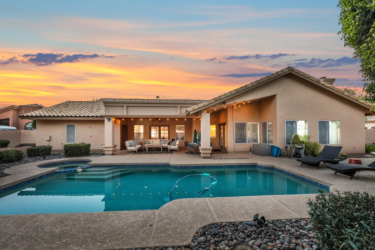A serene backyard scene at sunset featuring a sparkling swimming pool, inviting outdoor seating area, and a beautiful sky transitioning to dusk. The pool is the central focus, with ample space around it for relaxation and entertaining. The area is surrounded by lush landscaping, creating a private and tranquil atmosphere.