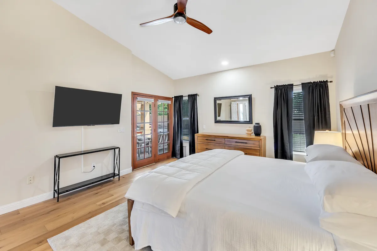 A tranquil bedroom with a plush bed, dressing table, large mirror, and French doors that open to the outside. A television sits opposite the bed, and soft natural light filters through, creating a restful atmosphere.