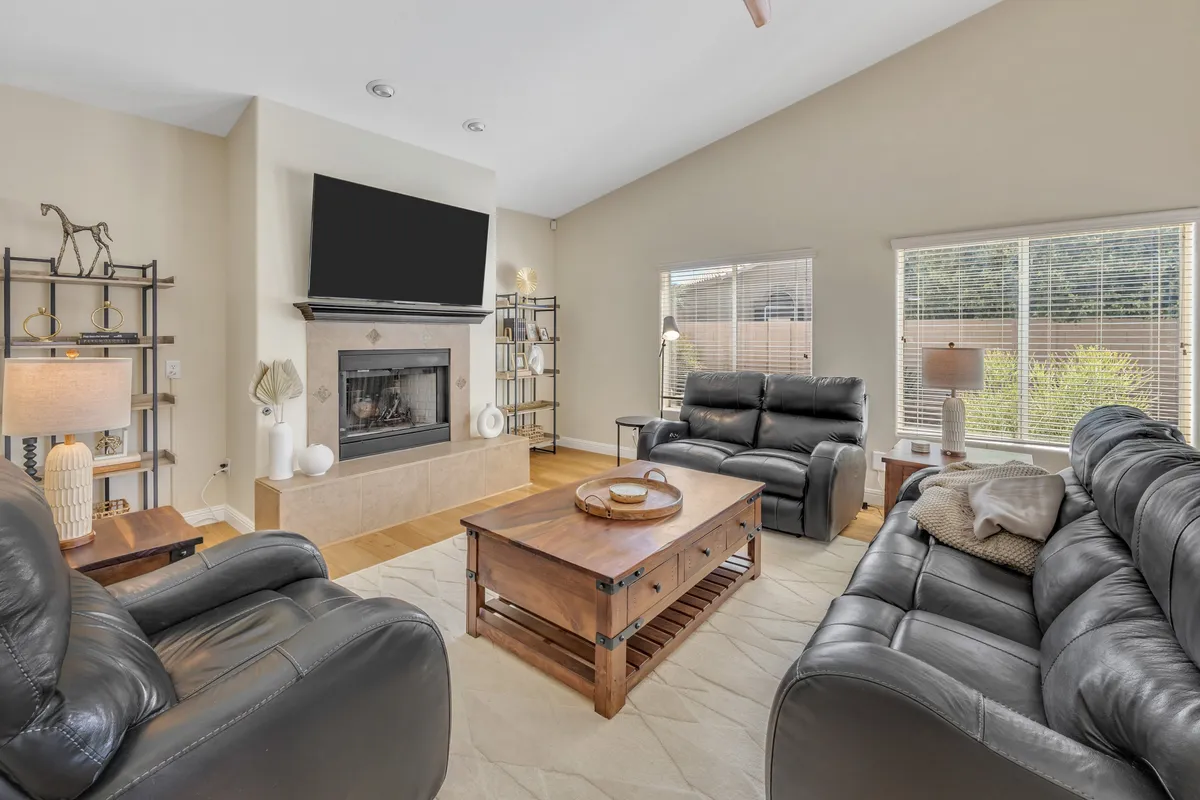 A warmly lit living room featuring a central fireplace flanked by sofas, a coffee table, and large windows that bathe the space in natural light. The arrangement invites both relaxation and conversation, with a television providing entertainment and the overall mood feeling cozy and welcoming.