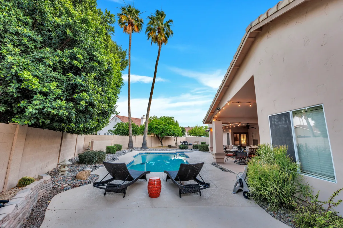 A sun-drenched backyard featuring a sparkling swimming pool surrounded by lounge chairs and palm trees. An outdoor dining area offers a perfect spot for meals or snacks after a day of adventure, while the overall scene promises fun, relaxation, and togetherness.