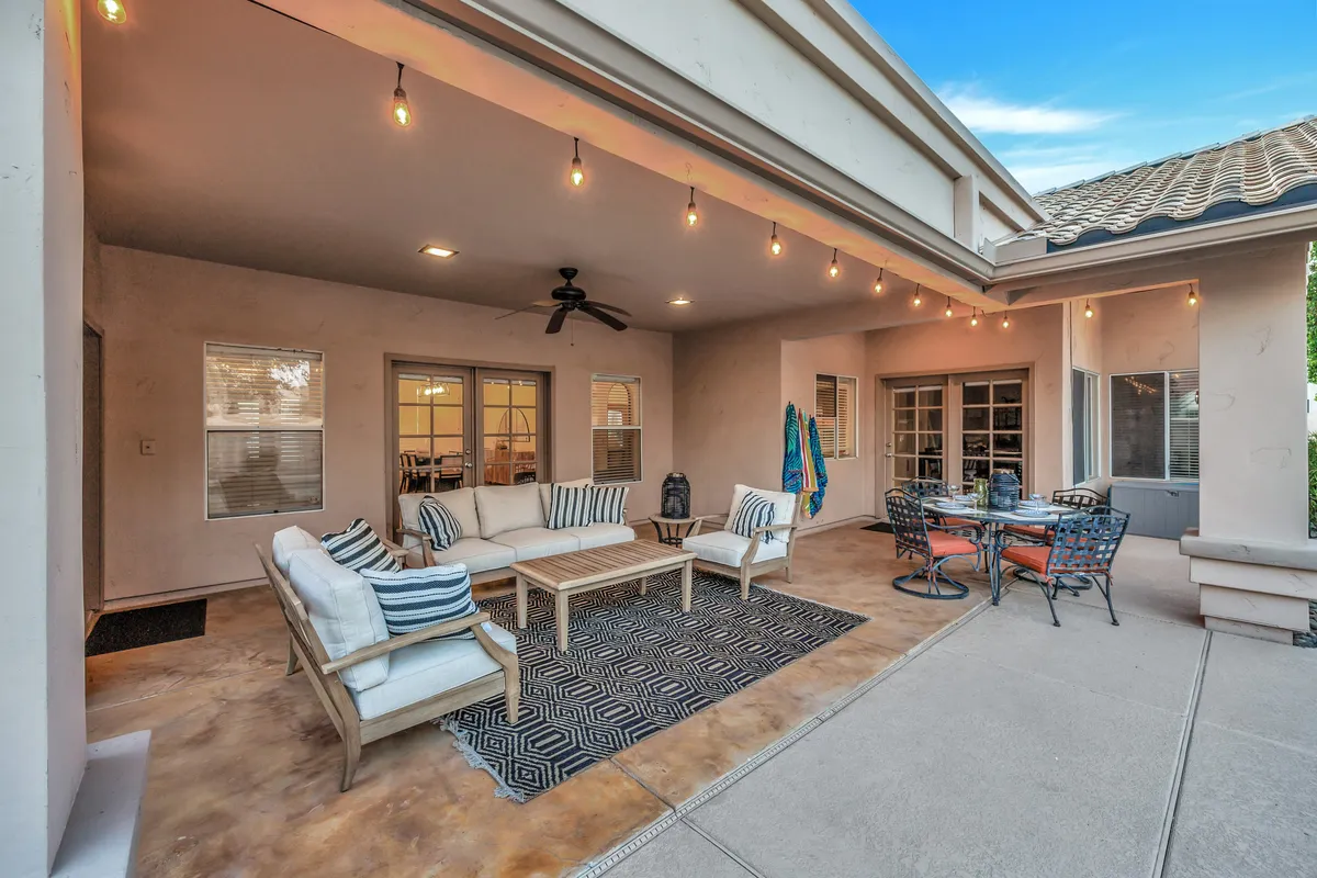 A stylish and cozy outdoor living room featuring a comfortable seating area, a chic dining set, a decorative rug, and strings of hanging lights overhead. The setup is ideal for both daytime lounging and evening gatherings under the stars, with plenty of space for socializing or quiet relaxation.